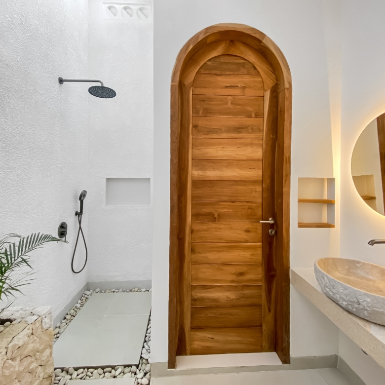Modern bathroom with a tall arched wooden door, a round backlit mirror, stone basin sinks, and a rainfall shower on the left.