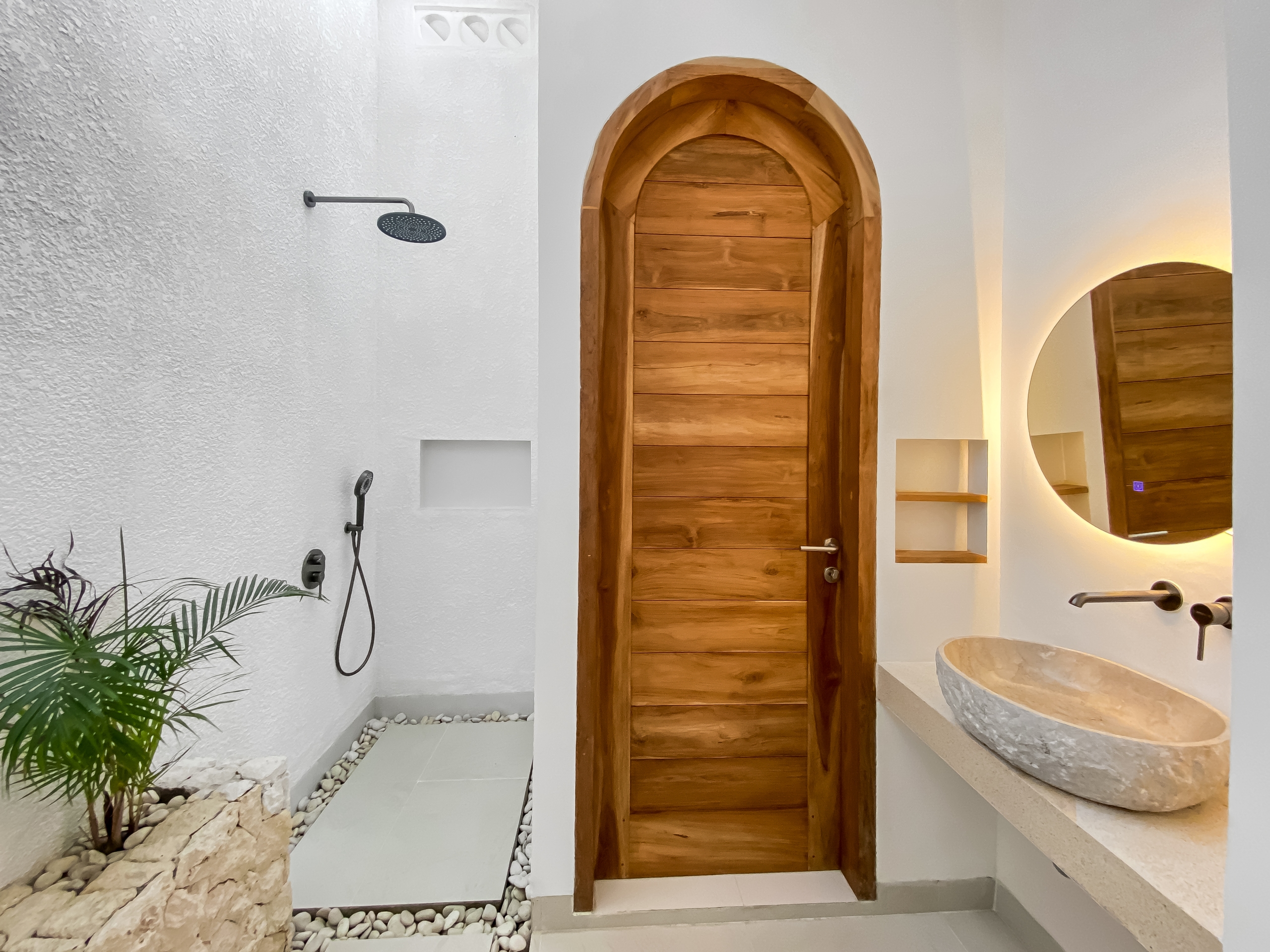 684e7ca2cca2a12fe47ca9cacc5cc052118914a3 Modern bathroom with a tall arched wooden door, a round backlit mirror, stone basin sinks, and a rainfall shower on the left.