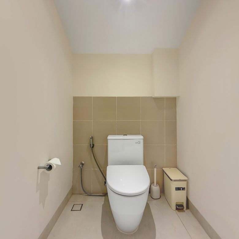 Narrow bathroom with beige walls and brown-tile backsplash, featuring a white toilet and a bidet spray hose to its left.