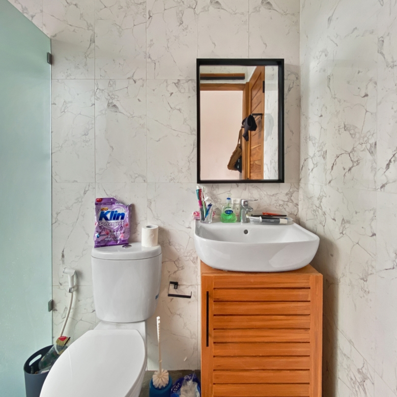 Marble-tiled bathroom with a white toilet, sink, wooden storage cabinet, and a wall mirror reflecting a wooden door.