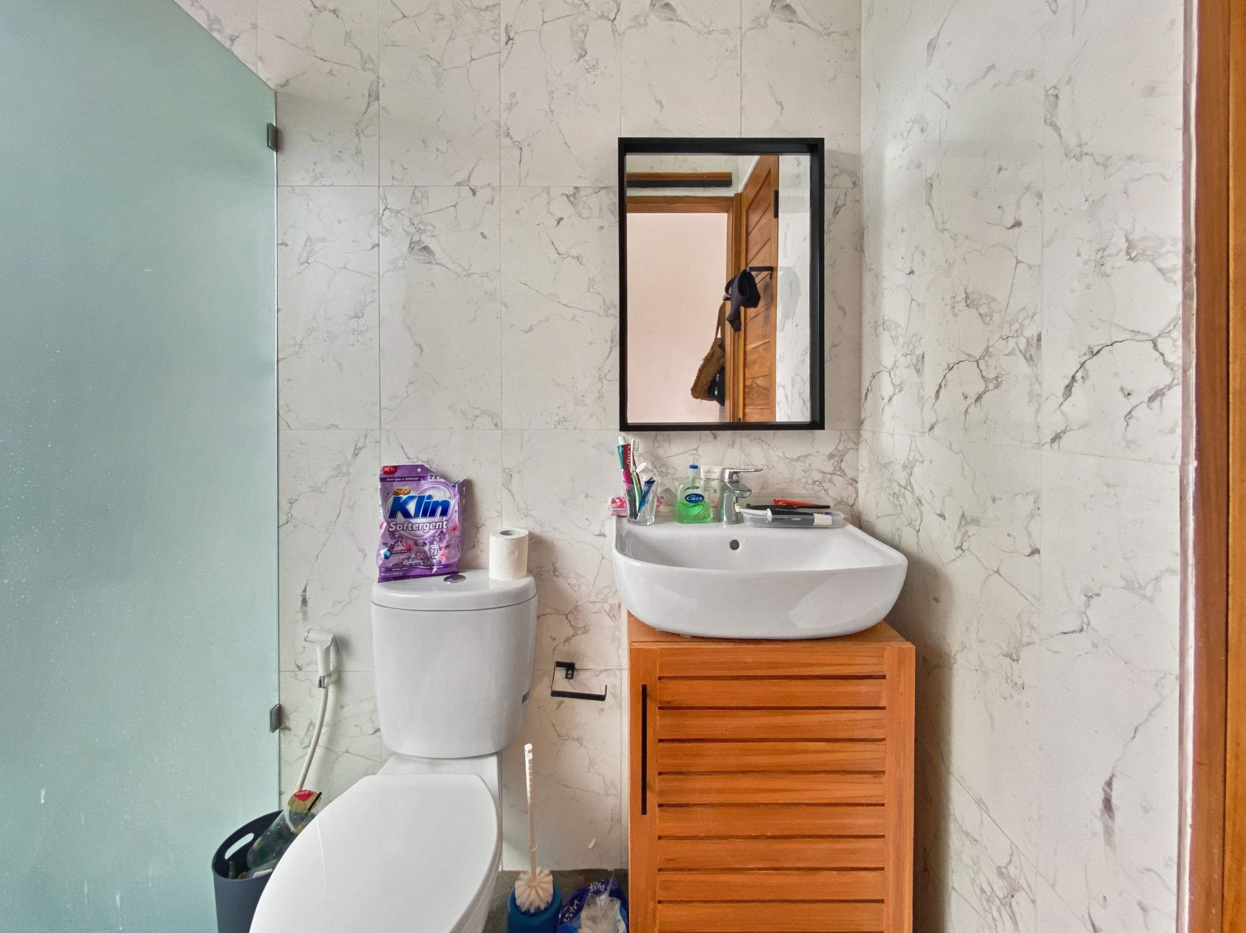6dc57823f652f02b786dcc903bd75f337bf136da Marble-tiled bathroom with a white toilet, sink, wooden storage cabinet, and a wall mirror reflecting a wooden door.