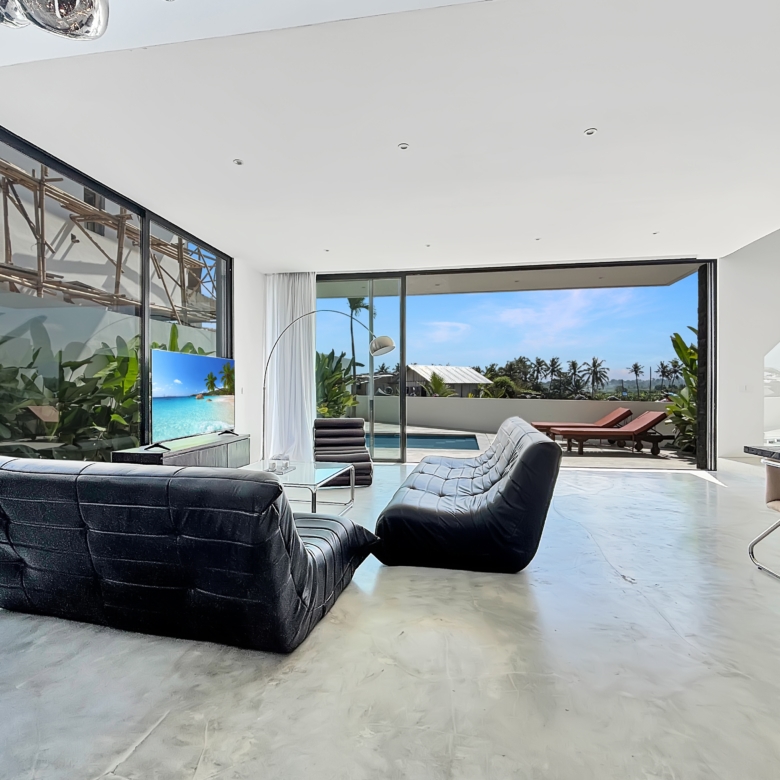 Open-plan living room with black curved leather sofa, glass coffee table, and floor-to-ceiling windows overlooking a pool and palm trees outside; bright, modern interior.