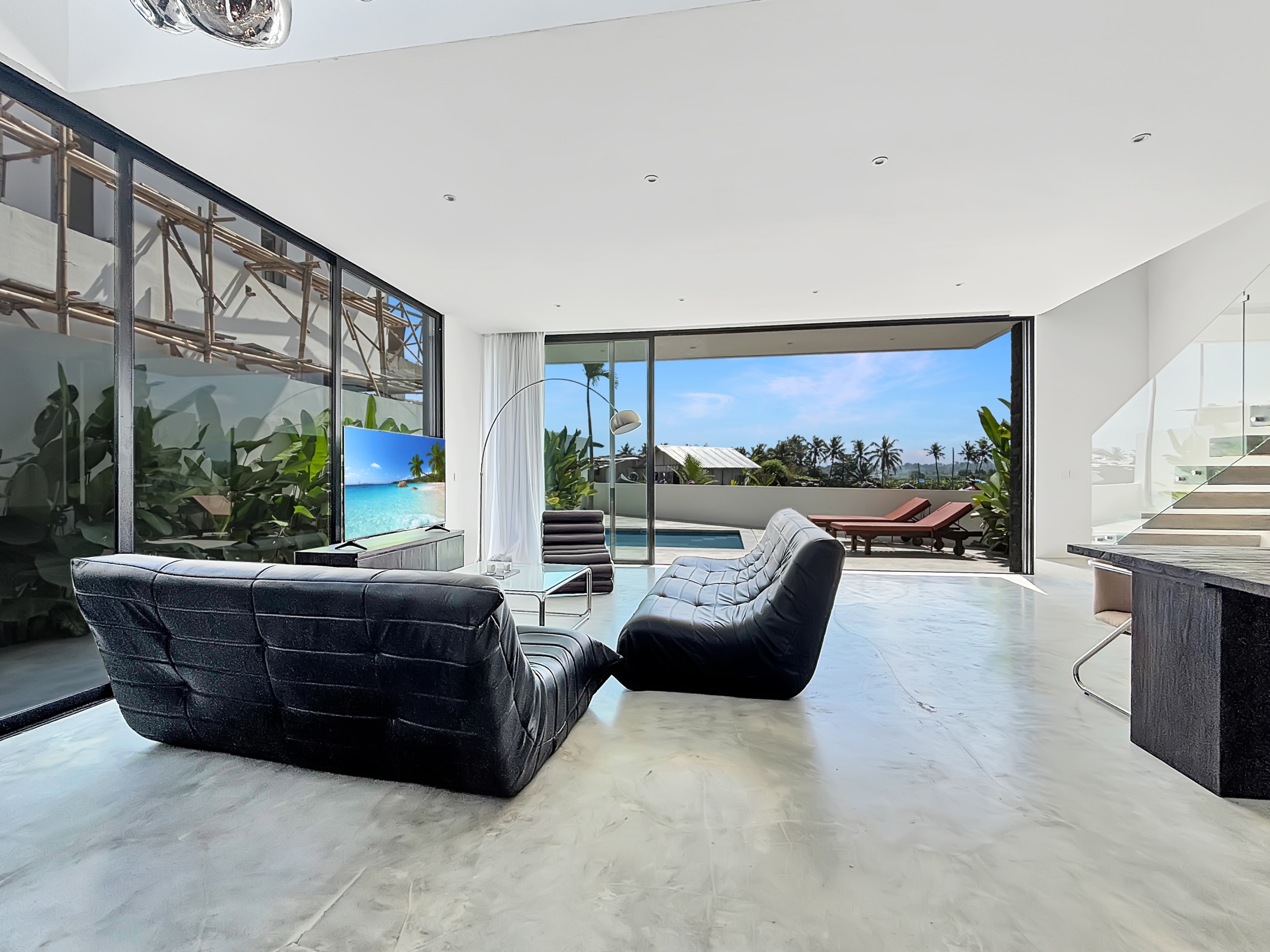 6e4784ffe55736ab8a22709665638f2a7123e044_6819-6a1d7bae378e1 Open-plan living room with black curved leather sofa, glass coffee table, and floor-to-ceiling windows overlooking a pool and palm trees outside; bright, modern interior.