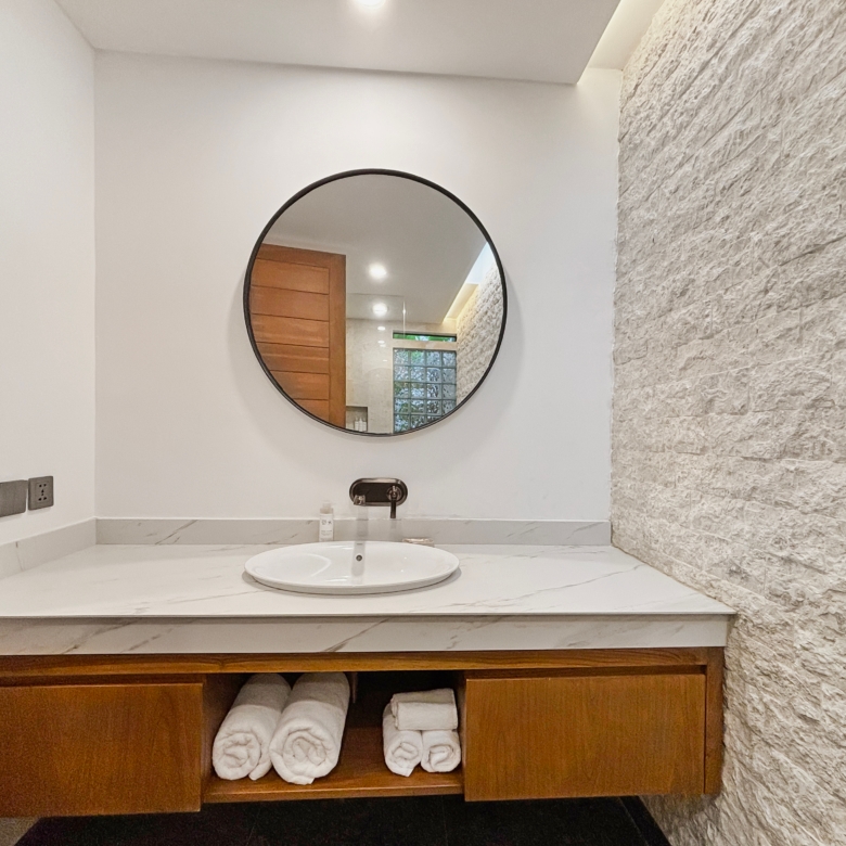 Bathroom vanity with a round mirror, marble countertop, and folded towels in the open shelf below.
