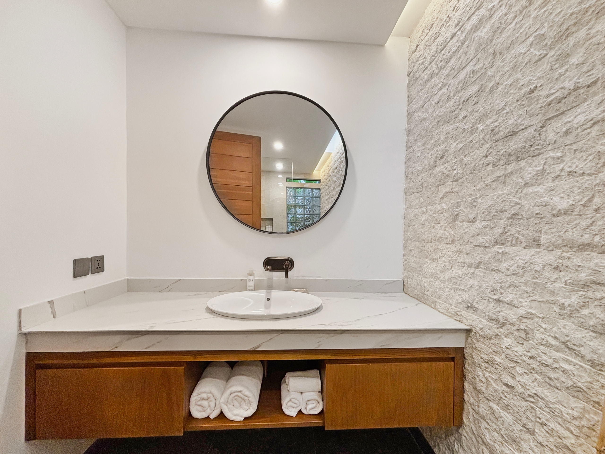 721360281529167101b1b6924266b774985314b9 Bathroom vanity with a round mirror, marble countertop, and folded towels in the open shelf below.