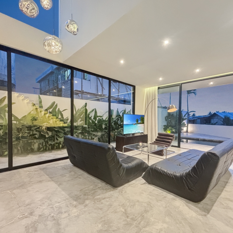 Modern living room with black leather chaise lounges, glass walls, and a TV showing a tropical beach scene; indoor plants nearby at dusk.