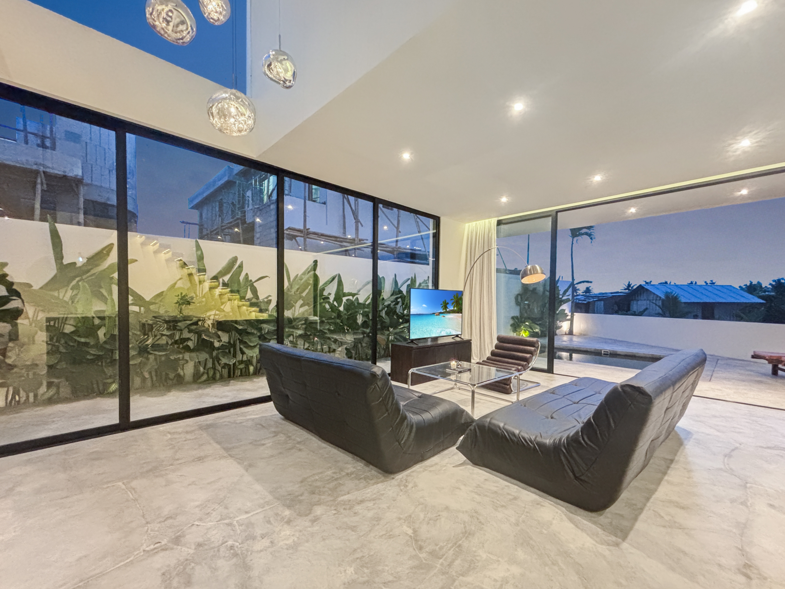 7286a35eece8ca5c2ec6f4746e1faafeb4284760_1010-6a05be0524e2c Modern living room with black leather chaise lounges, glass walls, and a TV showing a tropical beach scene; indoor plants nearby at dusk.