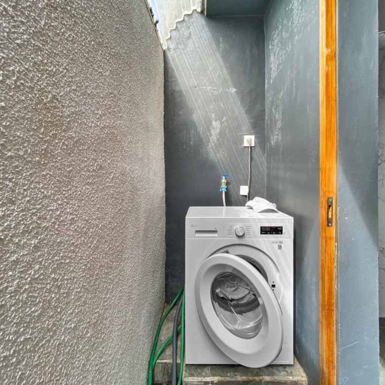 Washing machine in a narrow utility alcove with exposed hoses and an electrical outlet on a dark wall, textured concrete to the left and a wooden frame to the right, door ajar on green panels