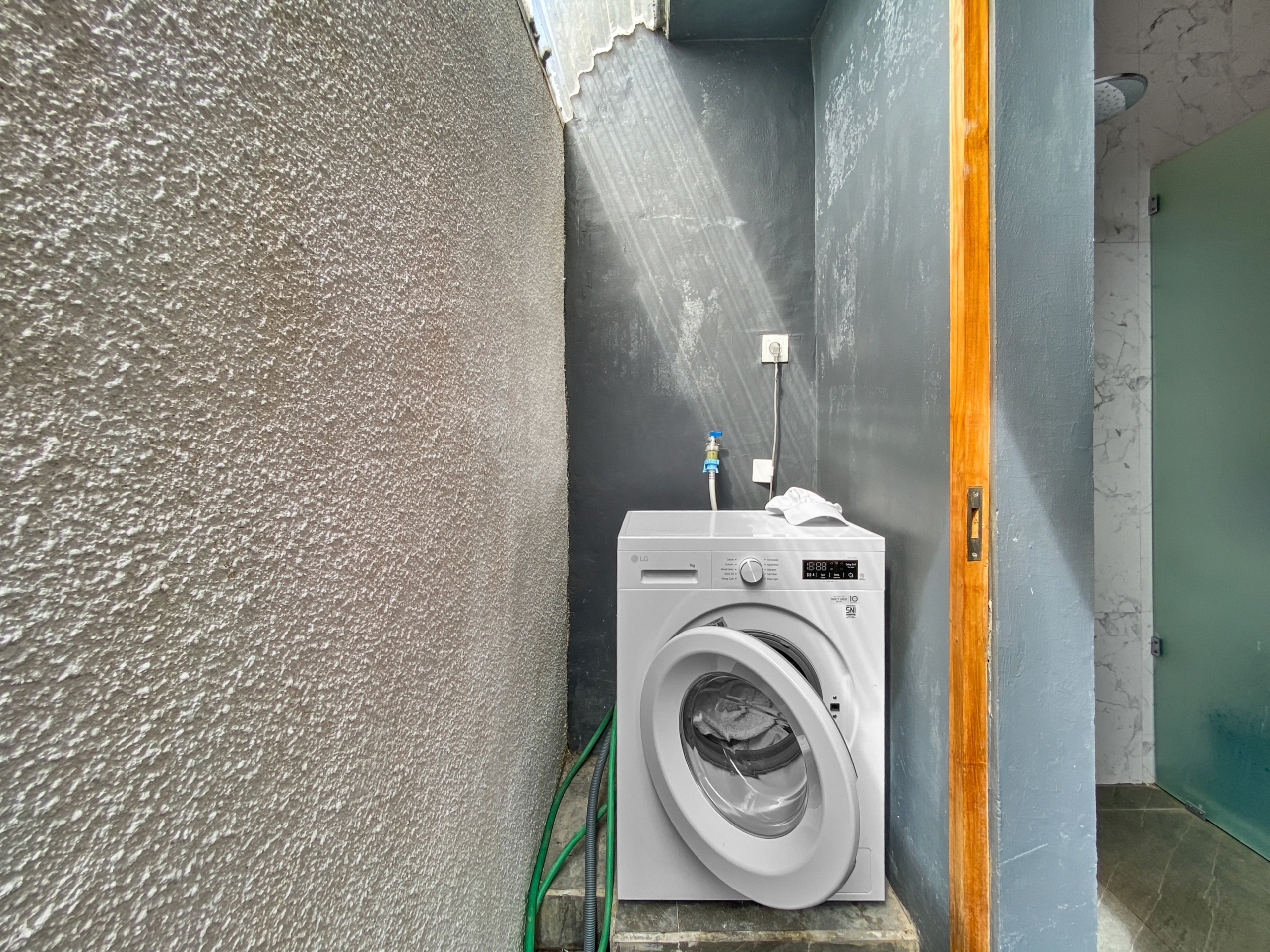 743682f2c32669487589bbfbf6182d93648986ab Washing machine in a narrow utility alcove with exposed hoses and an electrical outlet on a dark wall, textured concrete to the left and a wooden frame to the right, door ajar on green panels