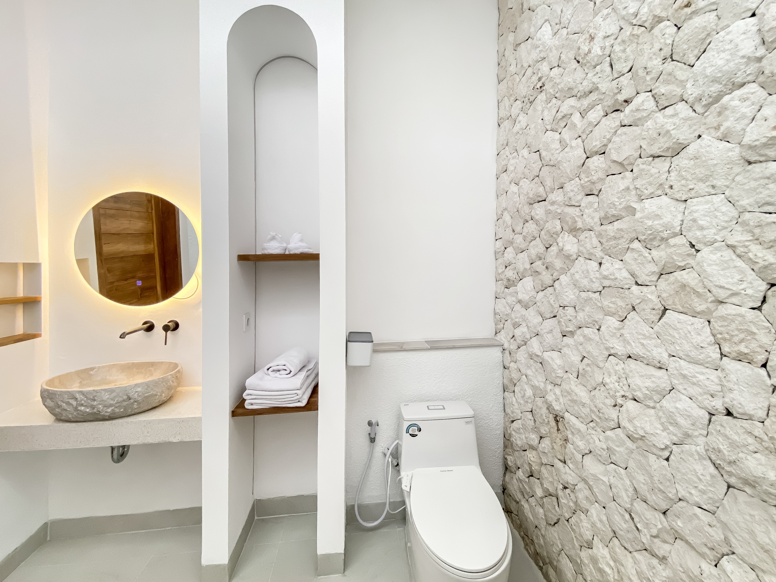 Modern bathroom interior featuring a round backlit mirror above a stone sink, white shelves, and a stacked stone wall on the right.] ,