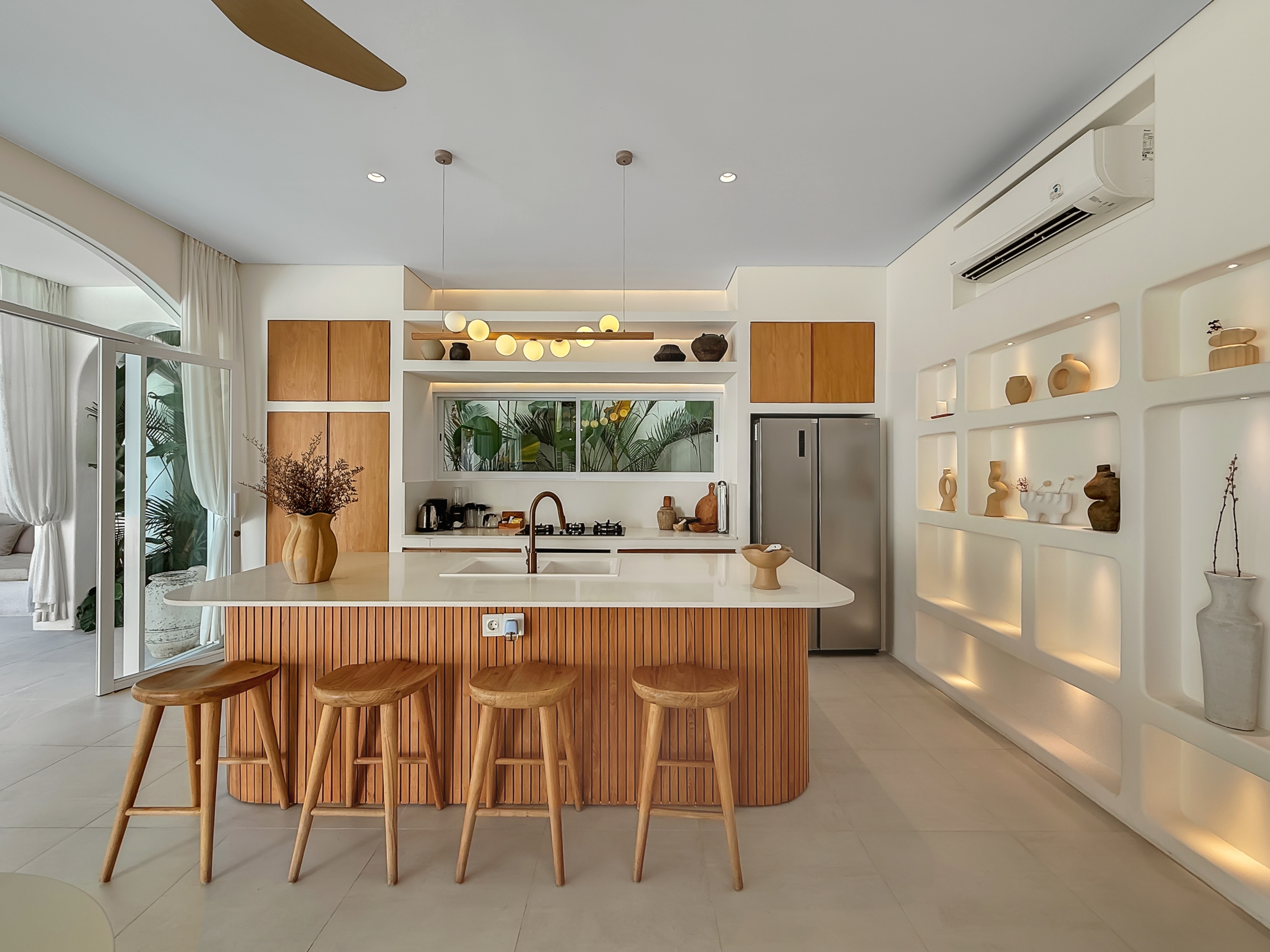 7c8a58e0429922b4ef4a5b4989de5827799e8071 Open-plan kitchen with a large wooden island and four wooden bar stools, white cabinets, and built-in shelves displaying pottery items.