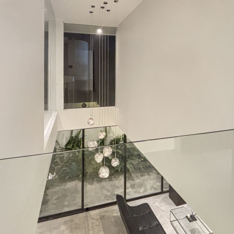 Modern stairwell/atrium with glass railing and hanging glass globe pendant lights above, plants visible below.