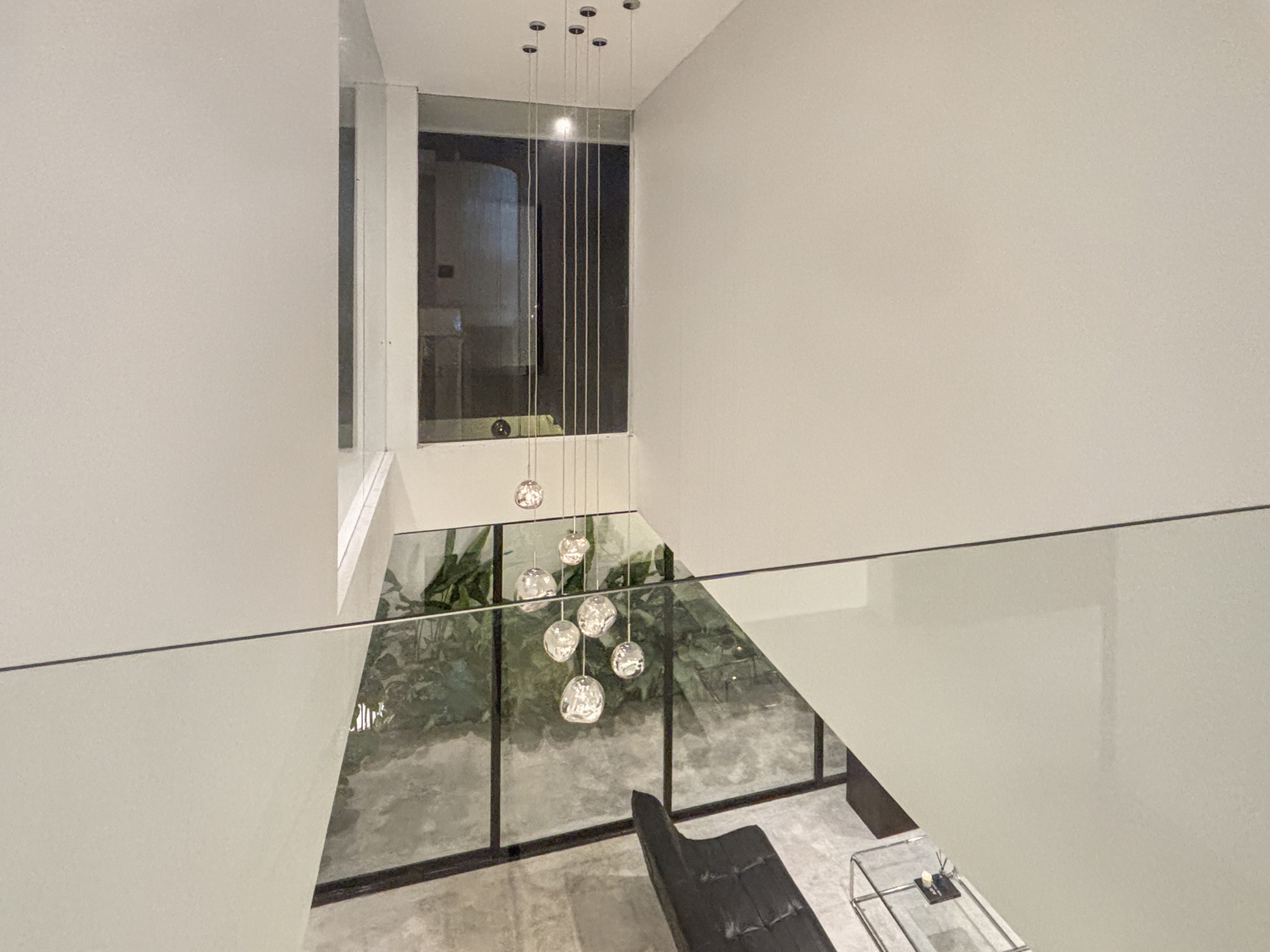 7cc221f6dbdf977ed96bc05010f1e85fbfa9d166_0896-6a05be075d617 Modern stairwell/atrium with glass railing and hanging glass globe pendant lights above, plants visible below.