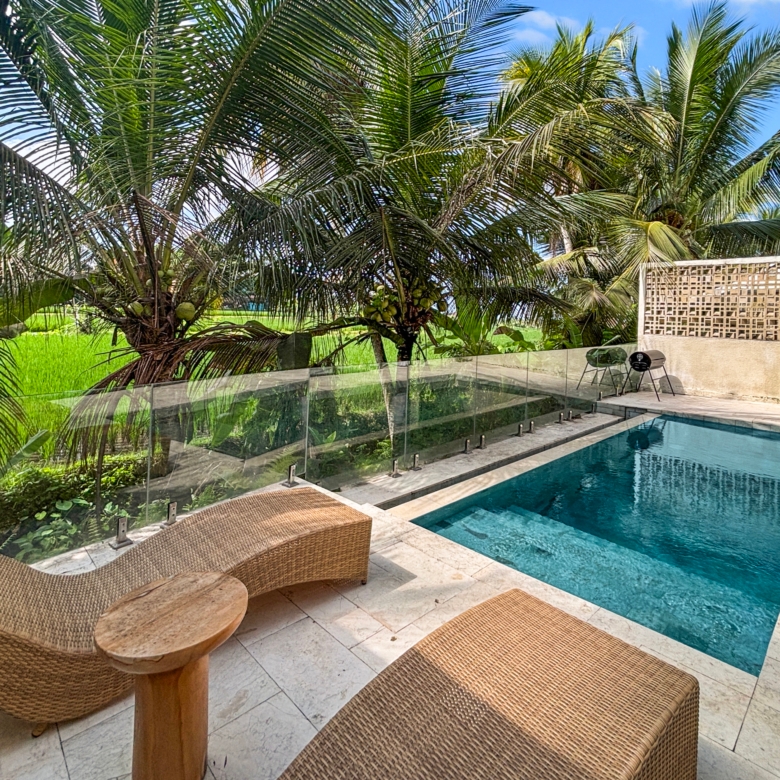 Poolside area with two wicker lounge chairs on a stone deck, glass railing, and lush palm trees under a blue sky.
