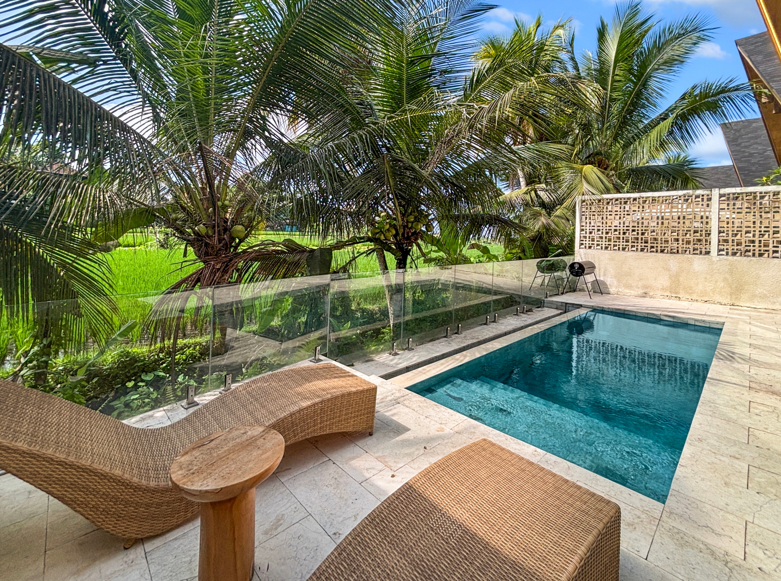 7fe726f38c027bf9536778af451169bcb5691ab2 Poolside area with two wicker lounge chairs on a stone deck, glass railing, and lush palm trees under a blue sky.