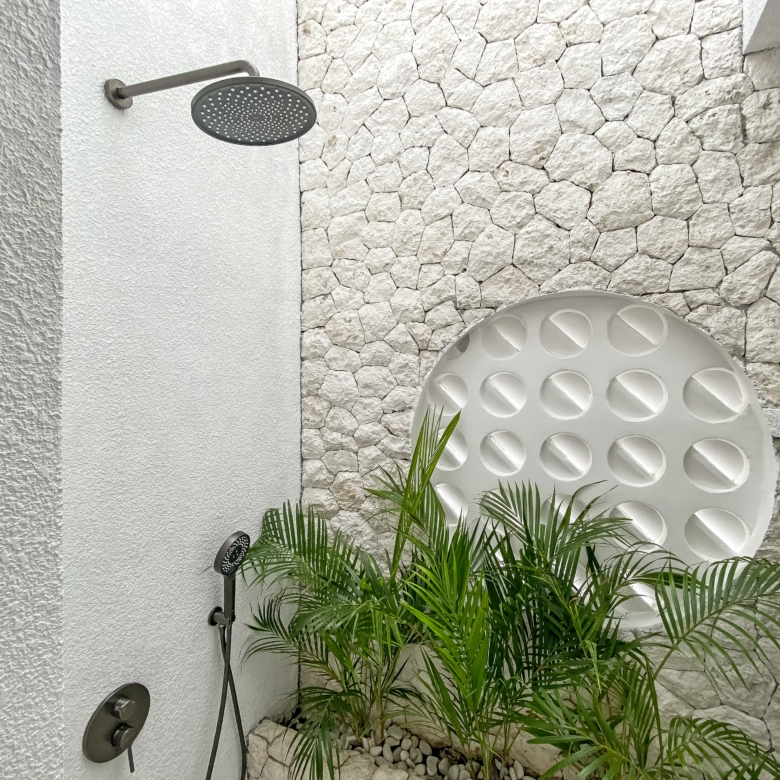 Outdoor shower with round rainfall head on a white wall, stone backdrop, pebbled base, and tropical plants nearby.
