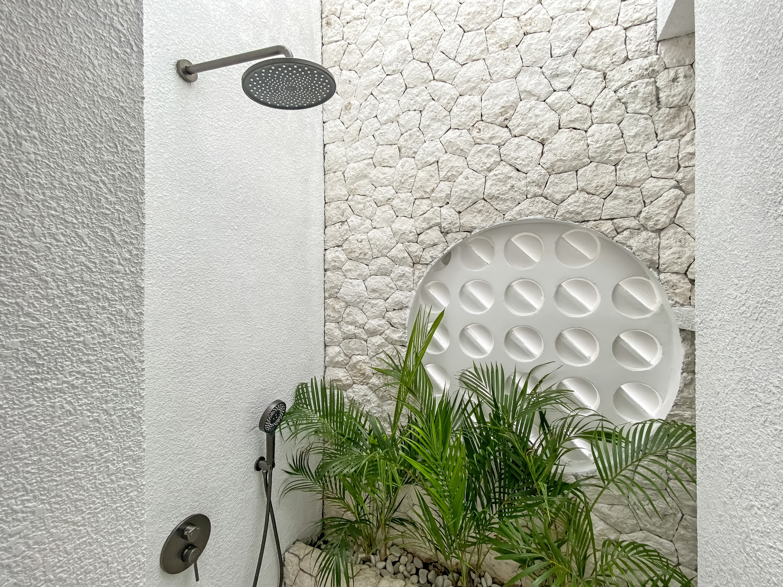 80215cd31abc50d0d78c9f0eb4609d34074bf2b5 Outdoor shower with round rainfall head on a white wall, stone backdrop, pebbled base, and tropical plants nearby.