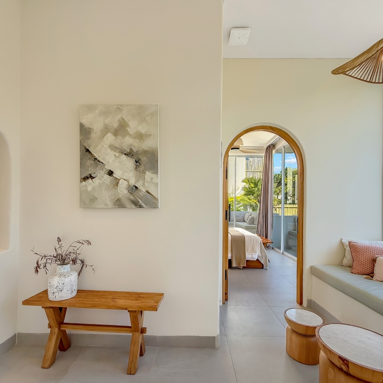 Interior of bright living area with arched doorway, wall art, and a wooden bench with a vase; doorway to a sunlit bedroom.