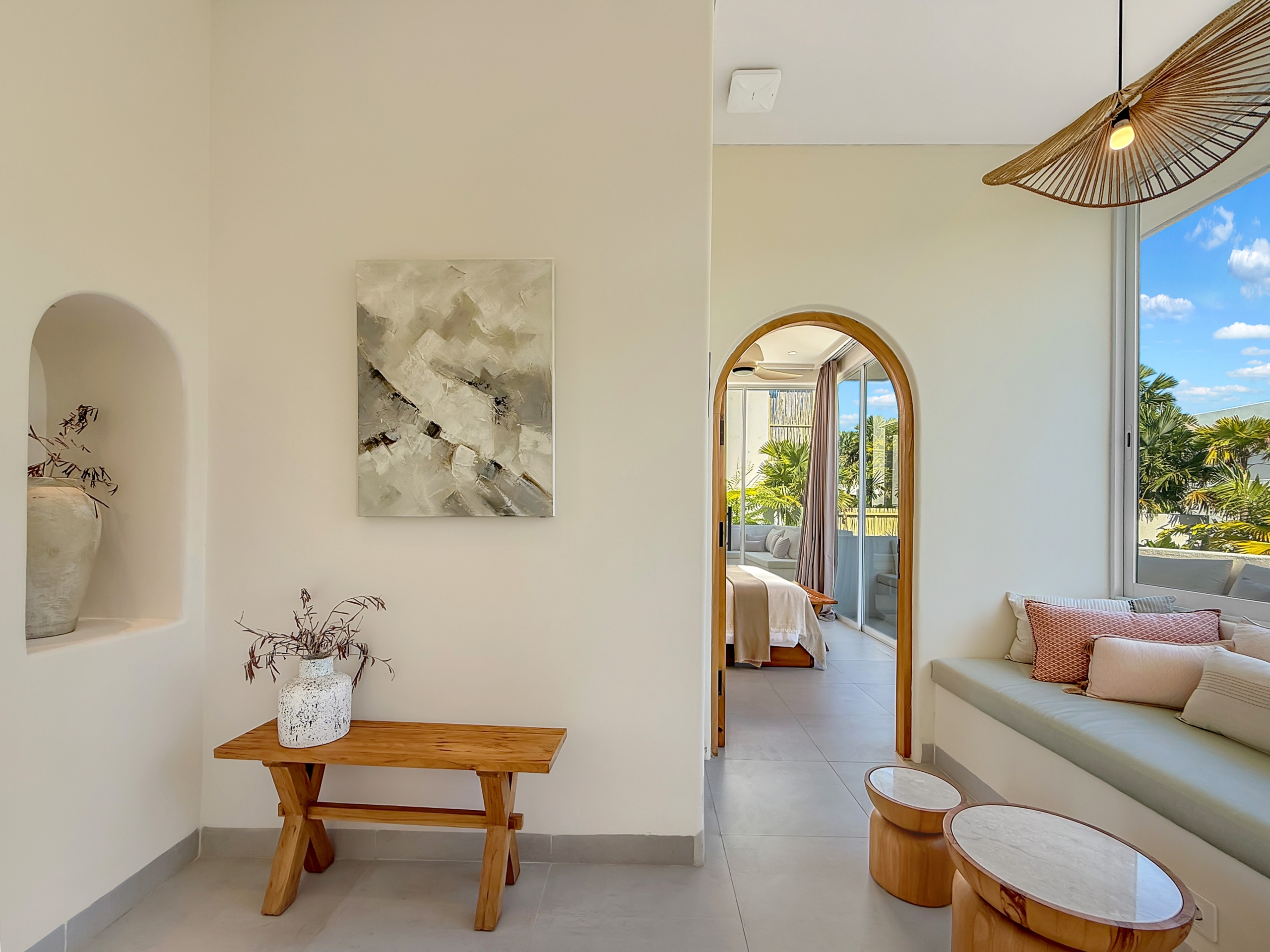 83940892018d89c1720312f86109c78aca9ed76f Interior of bright living area with arched doorway, wall art, and a wooden bench with a vase; doorway to a sunlit bedroom.