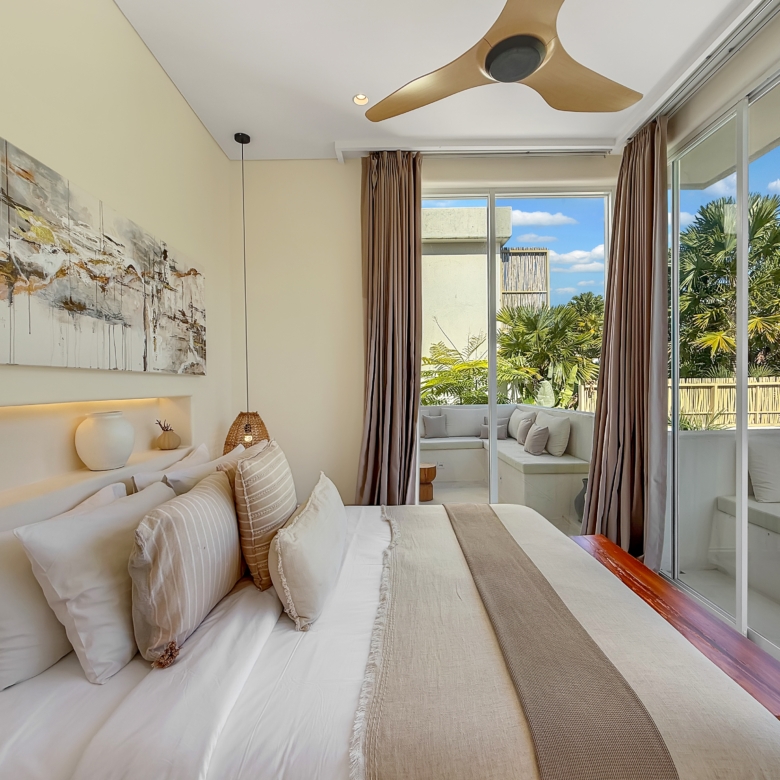 Bright hotel bedroom with white bedding, beige accents, and a glass wall opening to a patio with palm trees nearby.