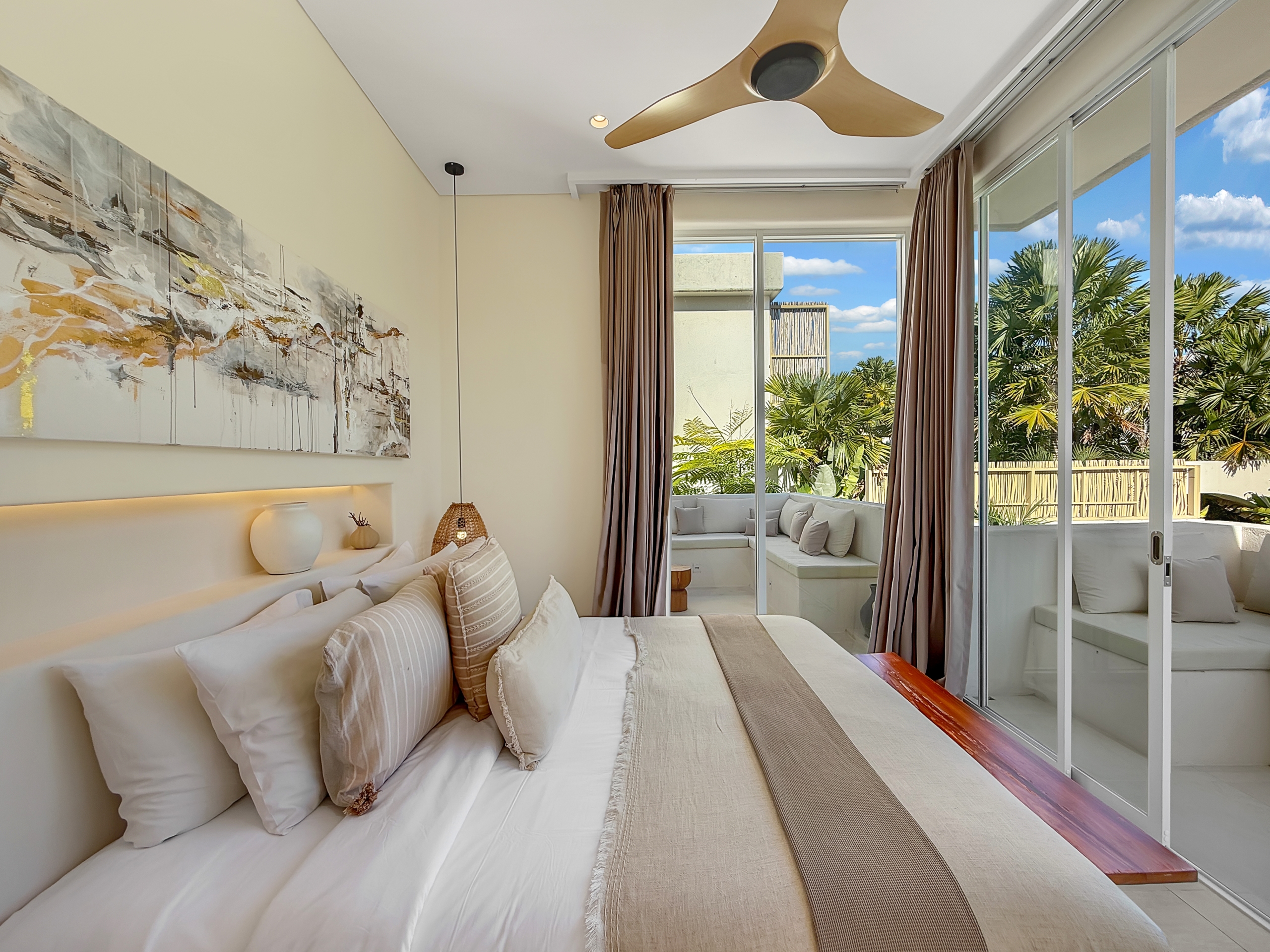 868462e37c8e68699e2e34cf445404af95bf4ec9 Bright hotel bedroom with white bedding, beige accents, and a glass wall opening to a patio with palm trees nearby.