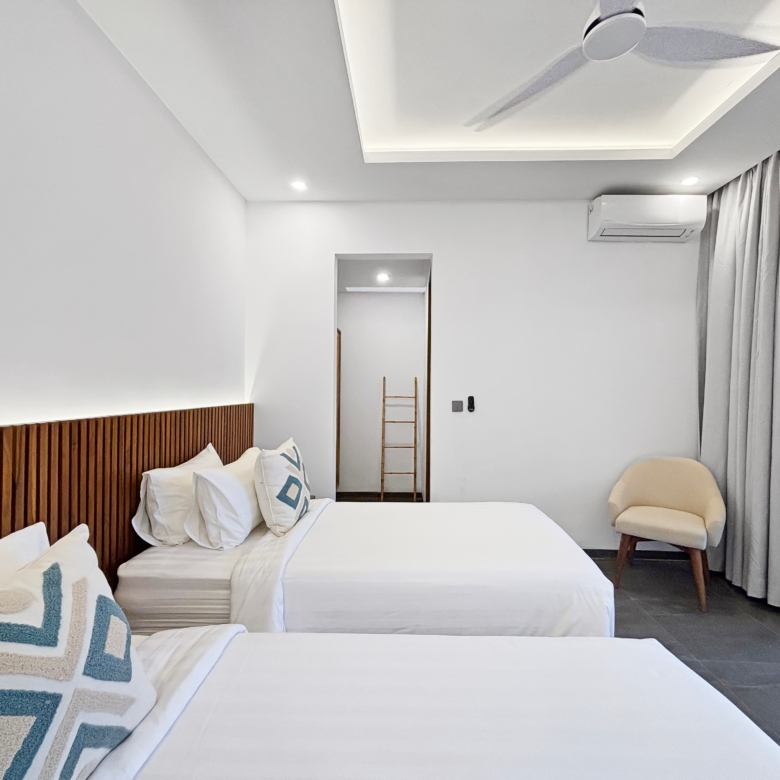 Hotel bedroom with two white beds, patterned pillows, and a wooden slat headboard under bright recessed lighting.