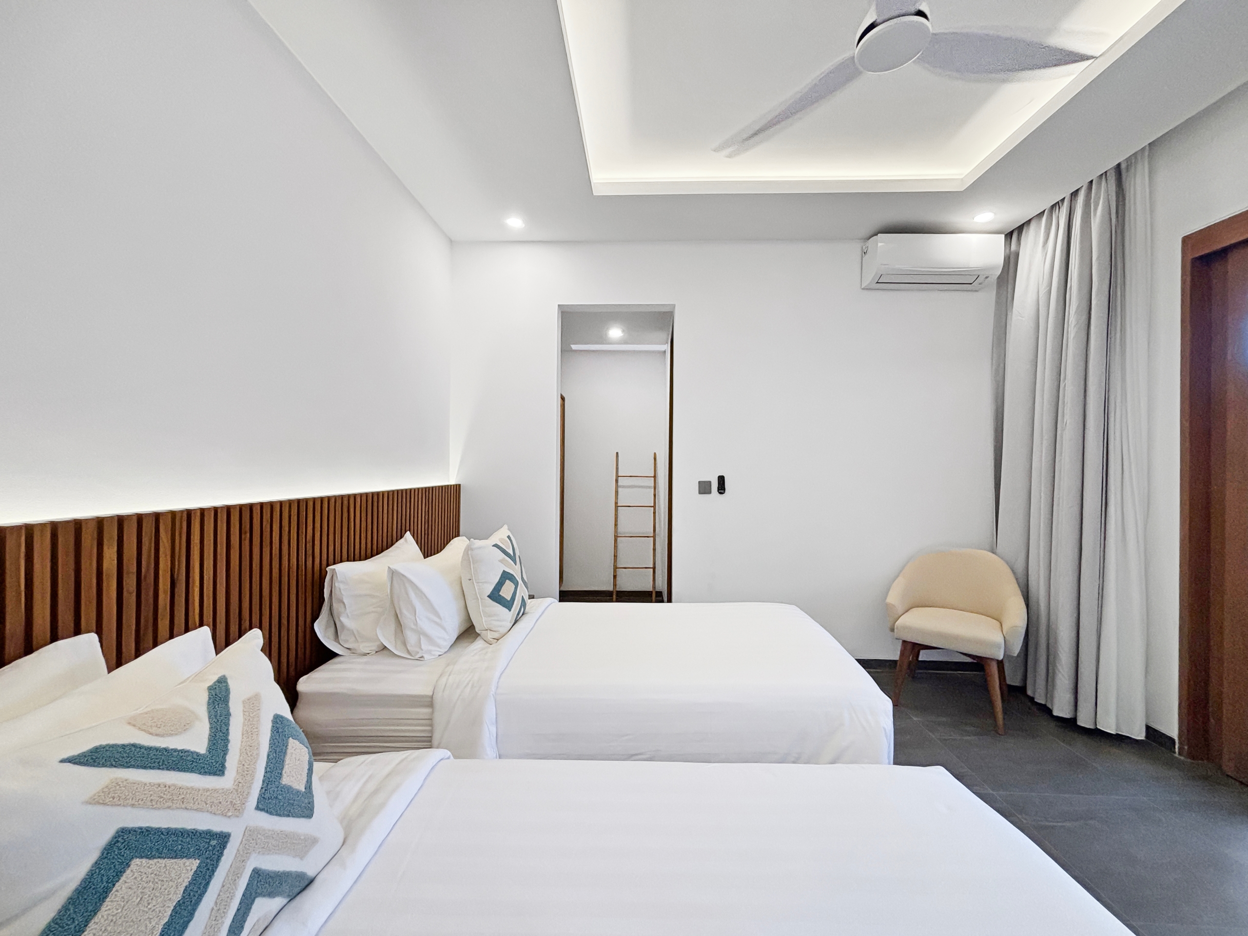 8be766f601ba4617e362b2dbdcb50f622b38cdd5 Hotel bedroom with two white beds, patterned pillows, and a wooden slat headboard under bright recessed lighting.