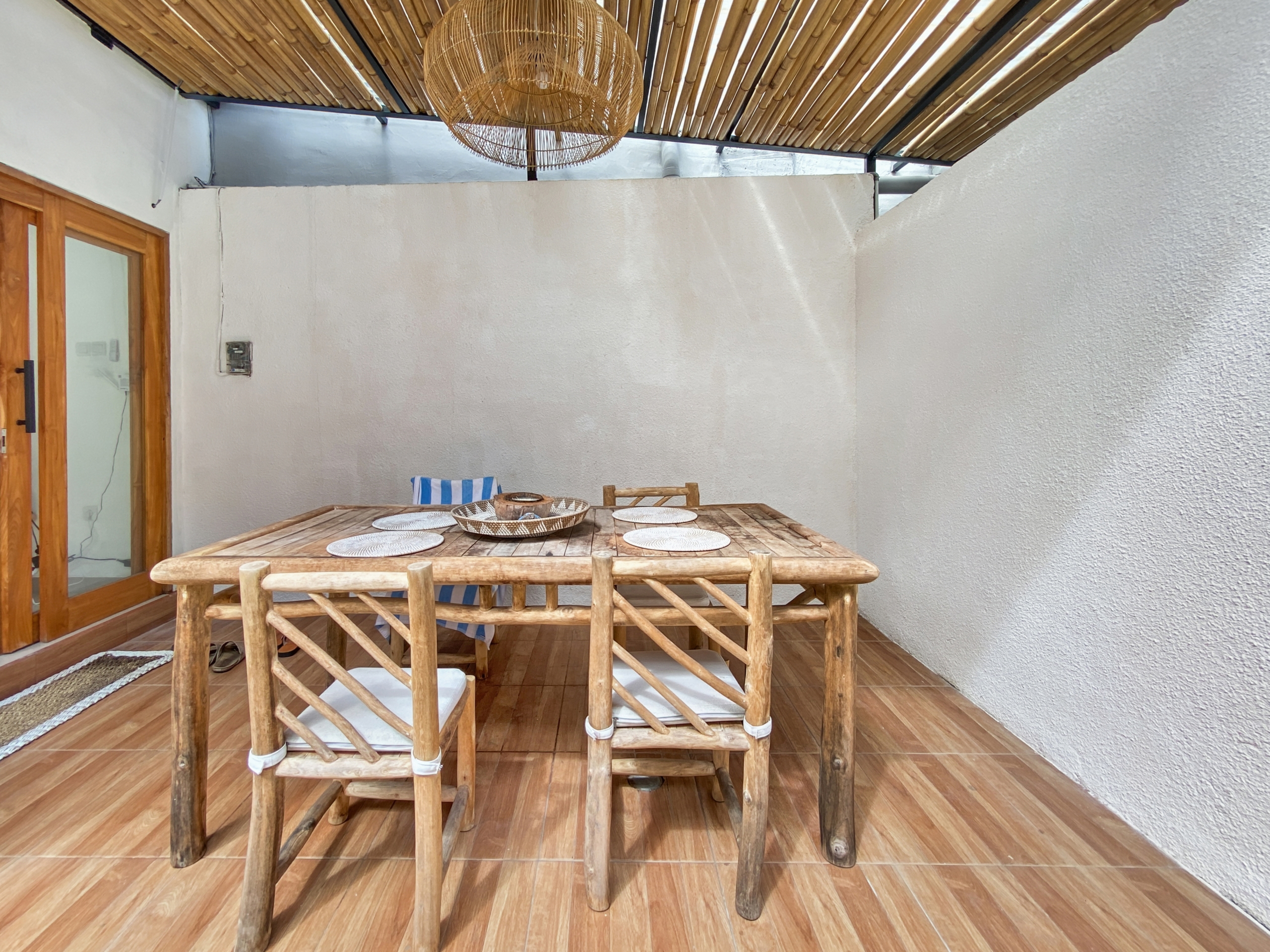 8d5a6cf06e2df6023350056ee7c314b4bf0f1156 Outdoor dining area with a rustic wooden table and matching chairs under a bamboo ceiling and wicker chandelier, on a tiled veranda.