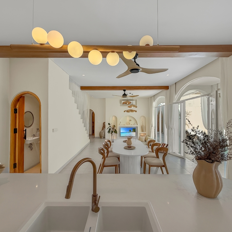 Open-plan kitchen and dining area with a white island, oval dining table, and wooden chairs under wooden ceiling beams and a ceiling fan.