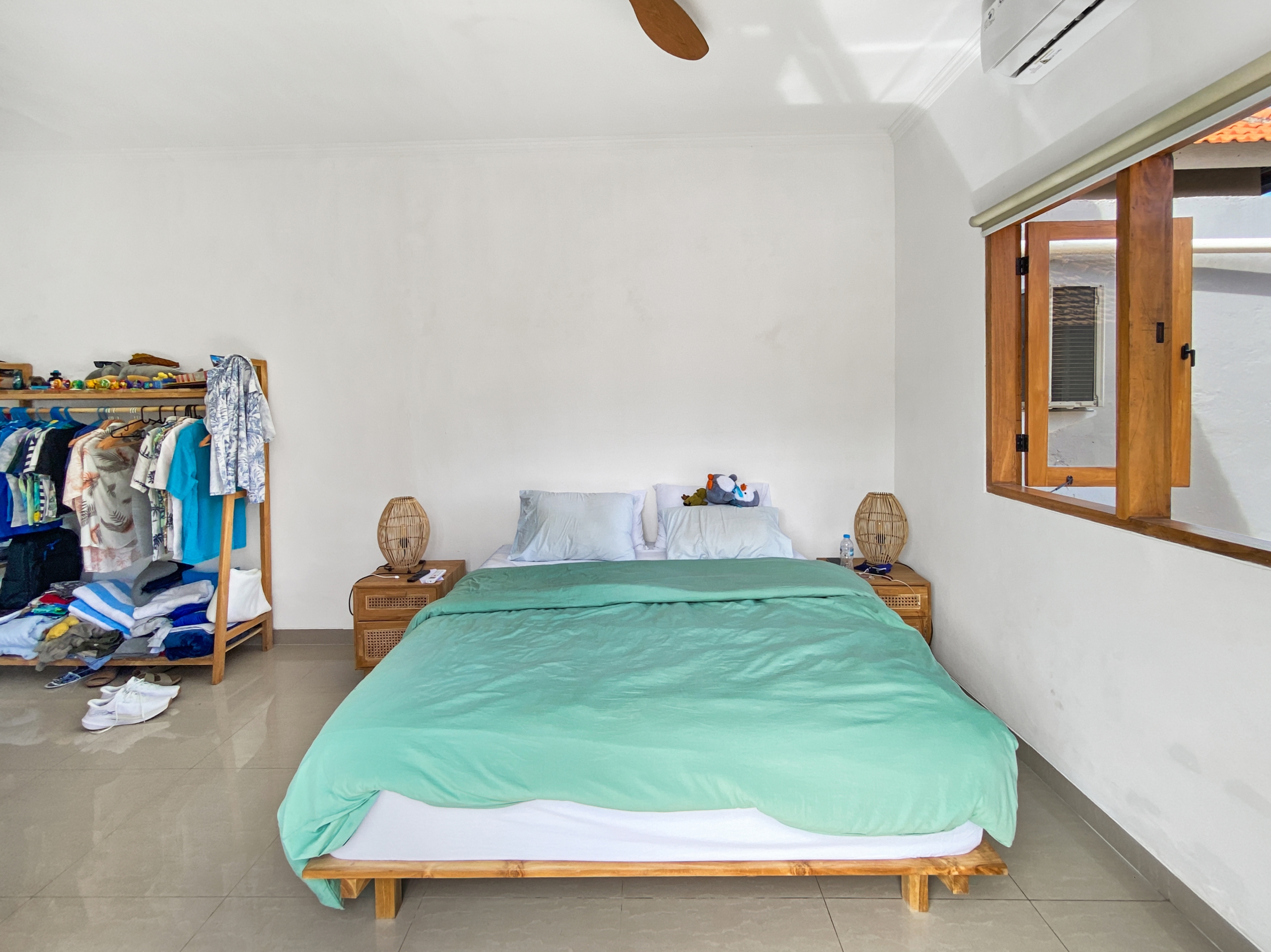 9455ba6c9f23c1c76db0444dc79a5671d9f21c94 Bedroom with a single bed in mint green bedding, a clothes rack on the left, and an open window on the right.