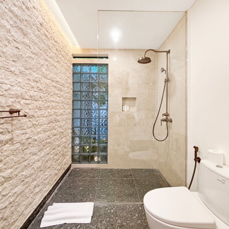 Bathroom with a textured stone accent wall, glass block window, and a rainfall shower; toilet and towels visible nearby.