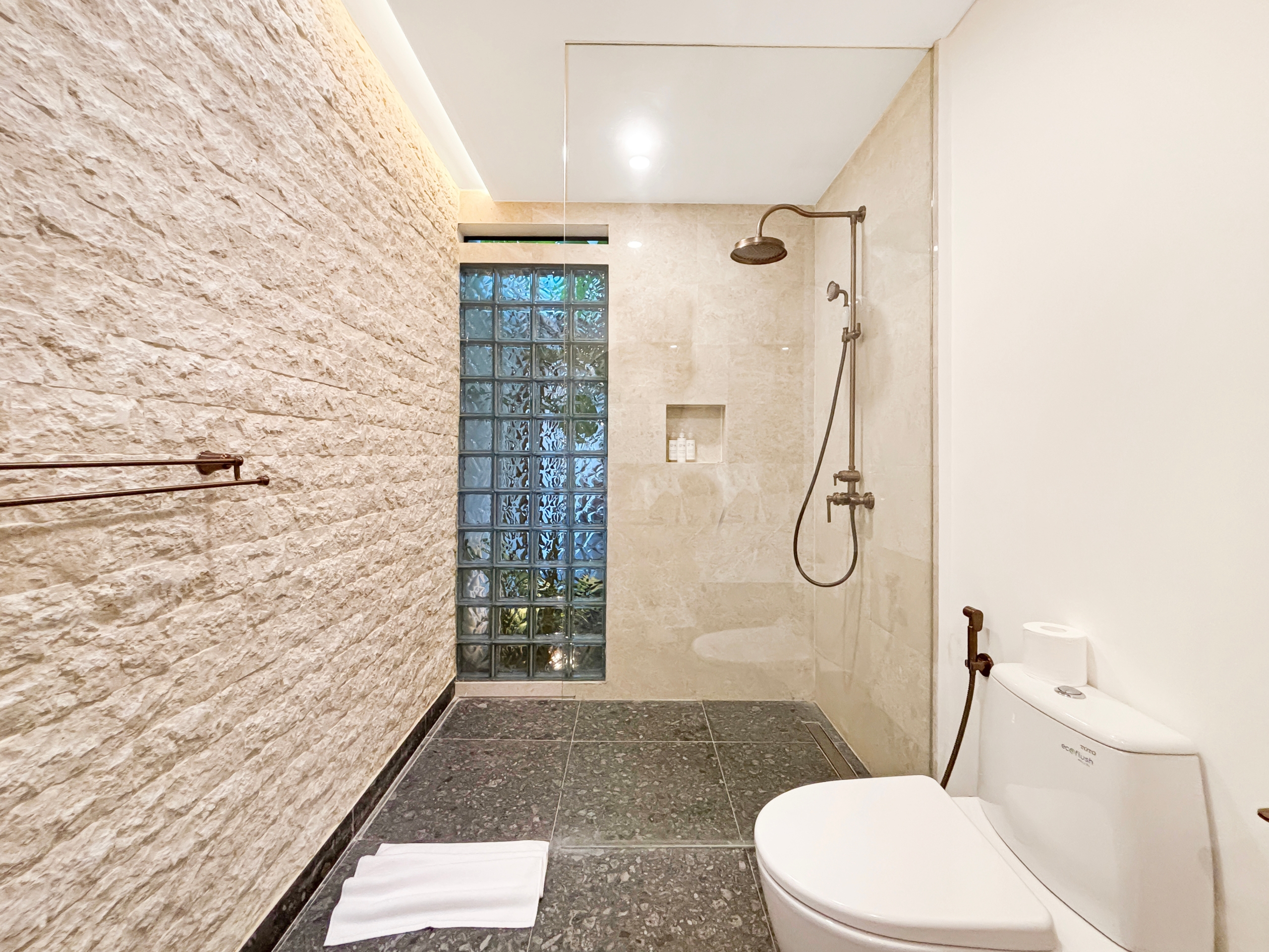 9b385359e3853a1a0ba84872fa90a4c879f8076d Bathroom with a textured stone accent wall, glass block window, and a rainfall shower; toilet and towels visible nearby.