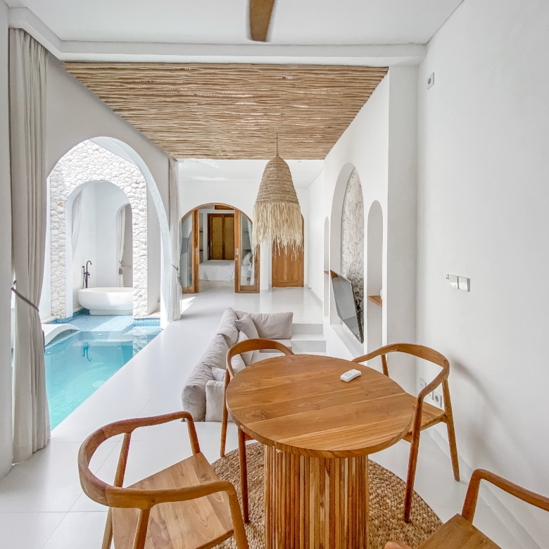 Bright white hallway with arches, a large woven pendant light, and a round wooden dining table with chairs; a pool is visible through the left arches.
