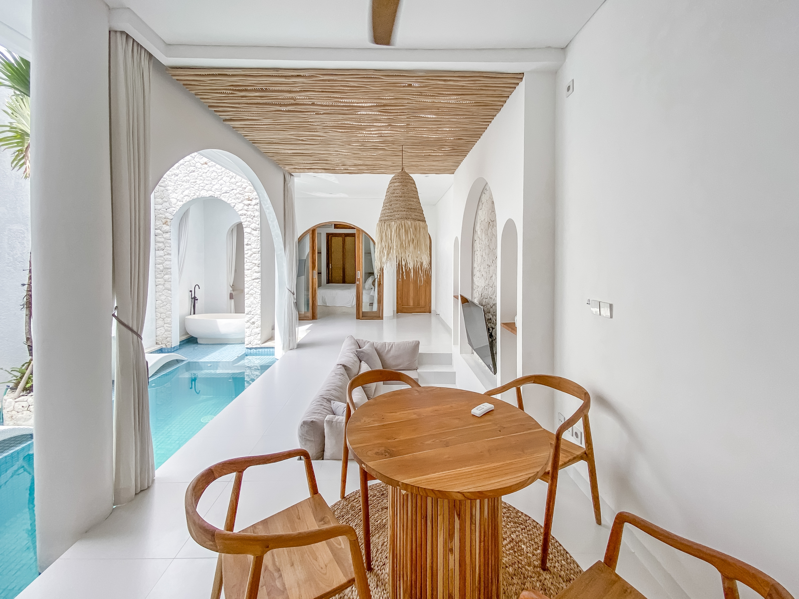 9e06c185f2d2c0b5f63c3456de3eff9d8f316c84 Bright white hallway with arches, a large woven pendant light, and a round wooden dining table with chairs; a pool is visible through the left arches.