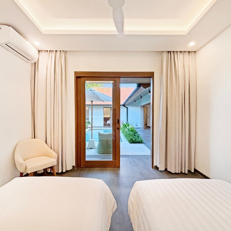 Hotel bedroom seen from between two white beds, with beige curtains, a wooden-framed glass door, and a patio outside with plants.