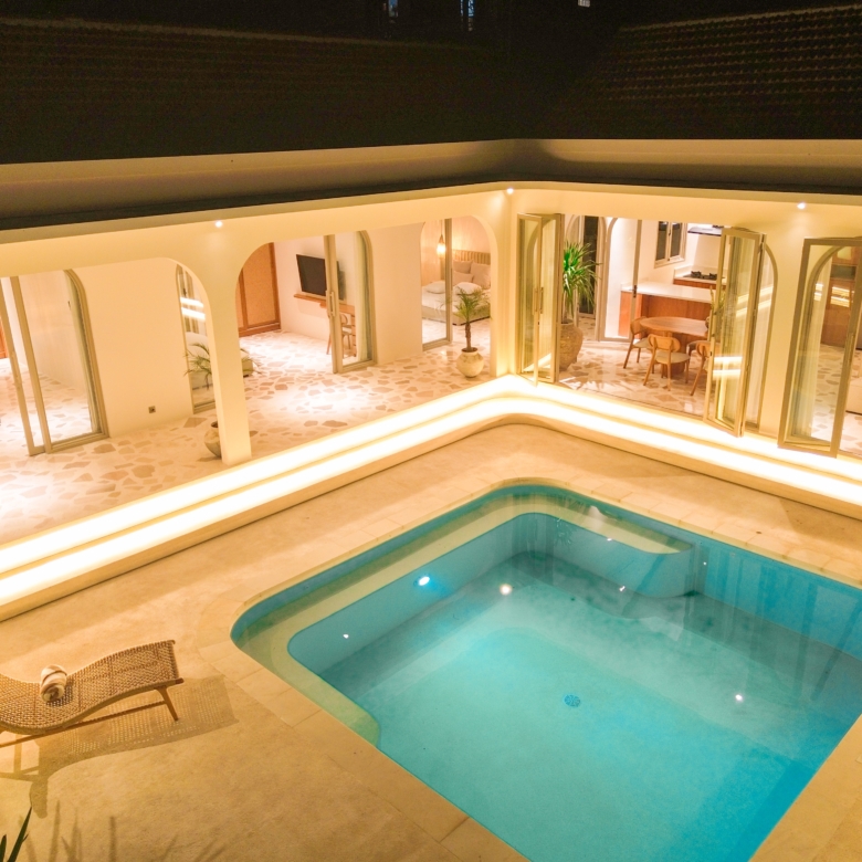 Indoor pool area at night with warm lighting, glass doors to living spaces, and potted plants surrounding the deck.
