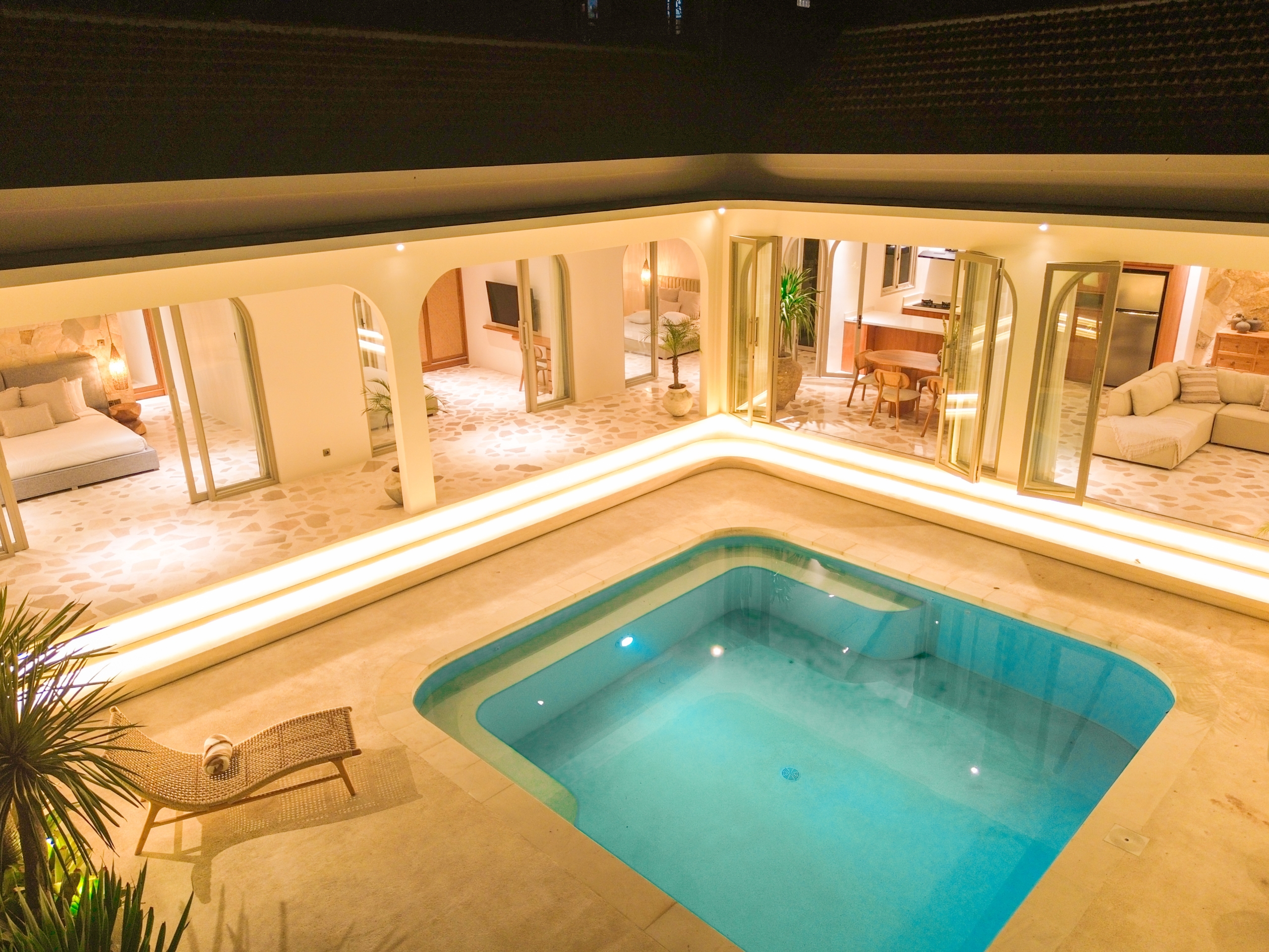 DJI_0140_4734-6a38c7b34bb52 Indoor pool area at night with warm lighting, glass doors to living spaces, and potted plants surrounding the deck.