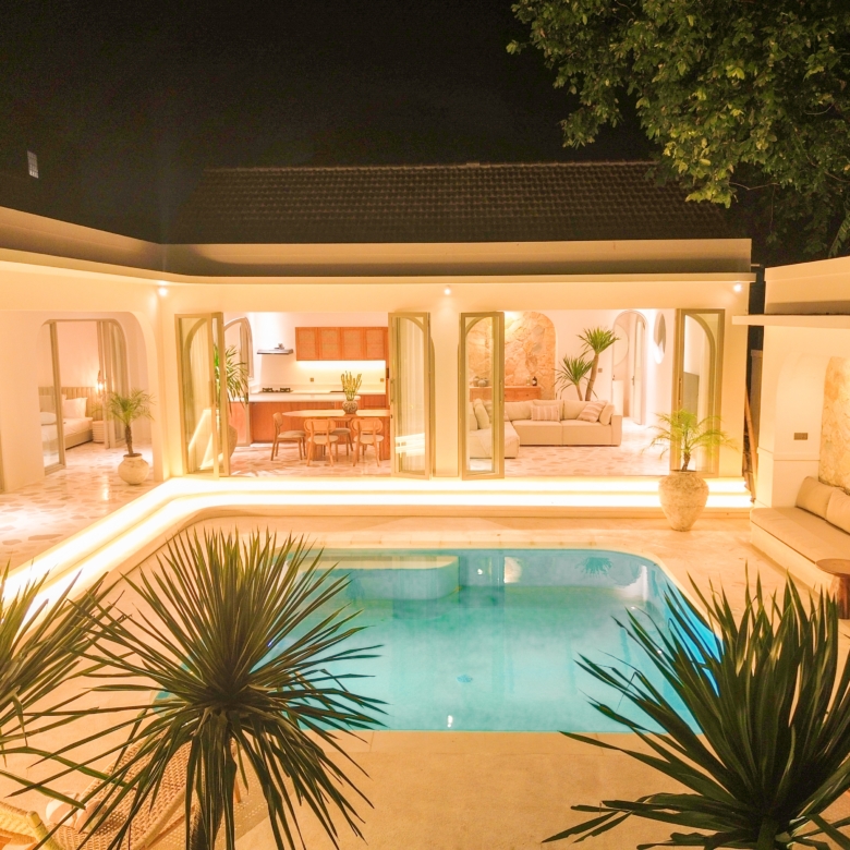 Nighttime view of a modern white villa with an illuminated open-plan living area and a turquoise pool in the foreground, tropical plants in the front.