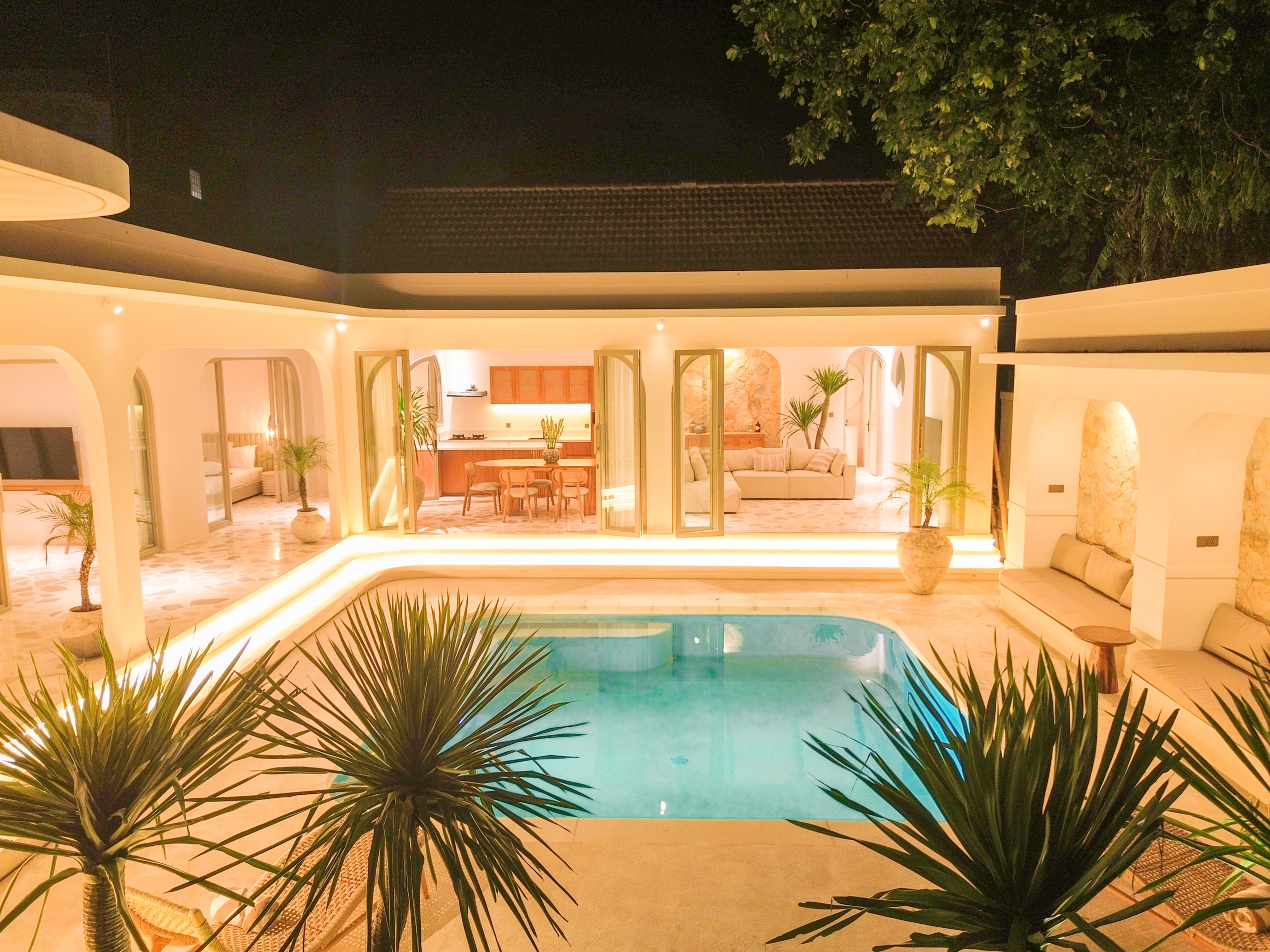 DJI_0143_4720-6a38c7b745cd7 Nighttime view of a modern white villa with an illuminated open-plan living area and a turquoise pool in the foreground, tropical plants in the front.