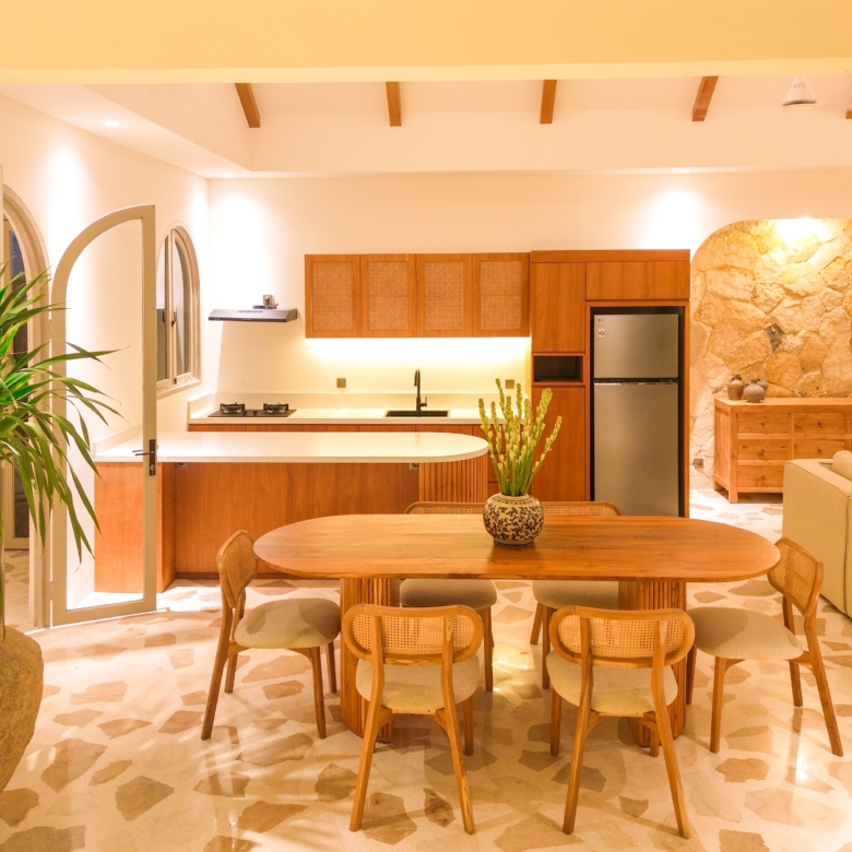 Open kitchen and dining area with a round wooden table, wicker chairs, wooden cabinets, and a stone accent wall to the right.