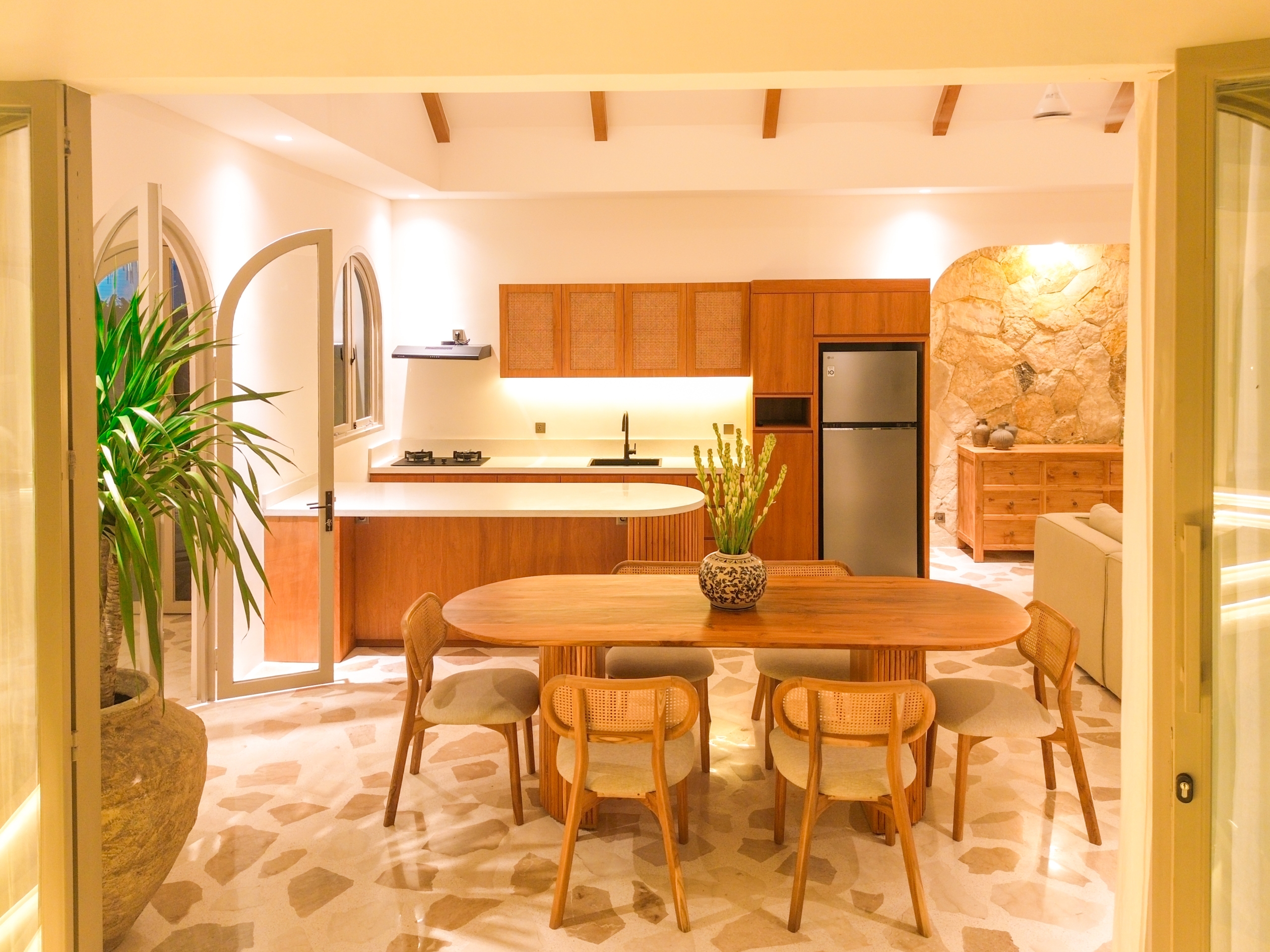 DJI_0166_4695-6a38c7b6c517e Open kitchen and dining area with a round wooden table, wicker chairs, wooden cabinets, and a stone accent wall to the right.