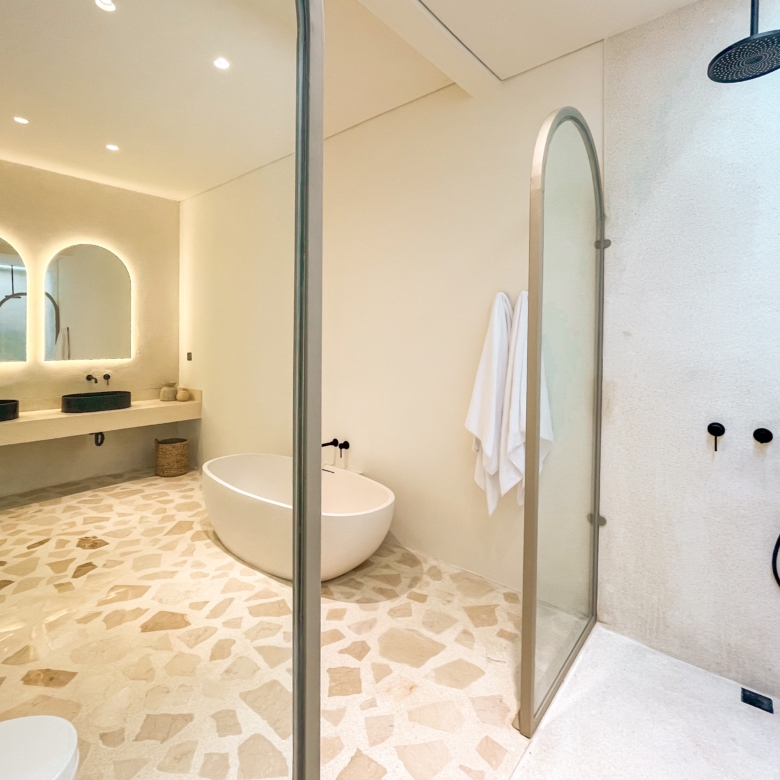 Modern bathroom with a glass shower enclosure, freestanding tub, and double sinks beneath backlit arched mirrors. Decorative pebble floor.