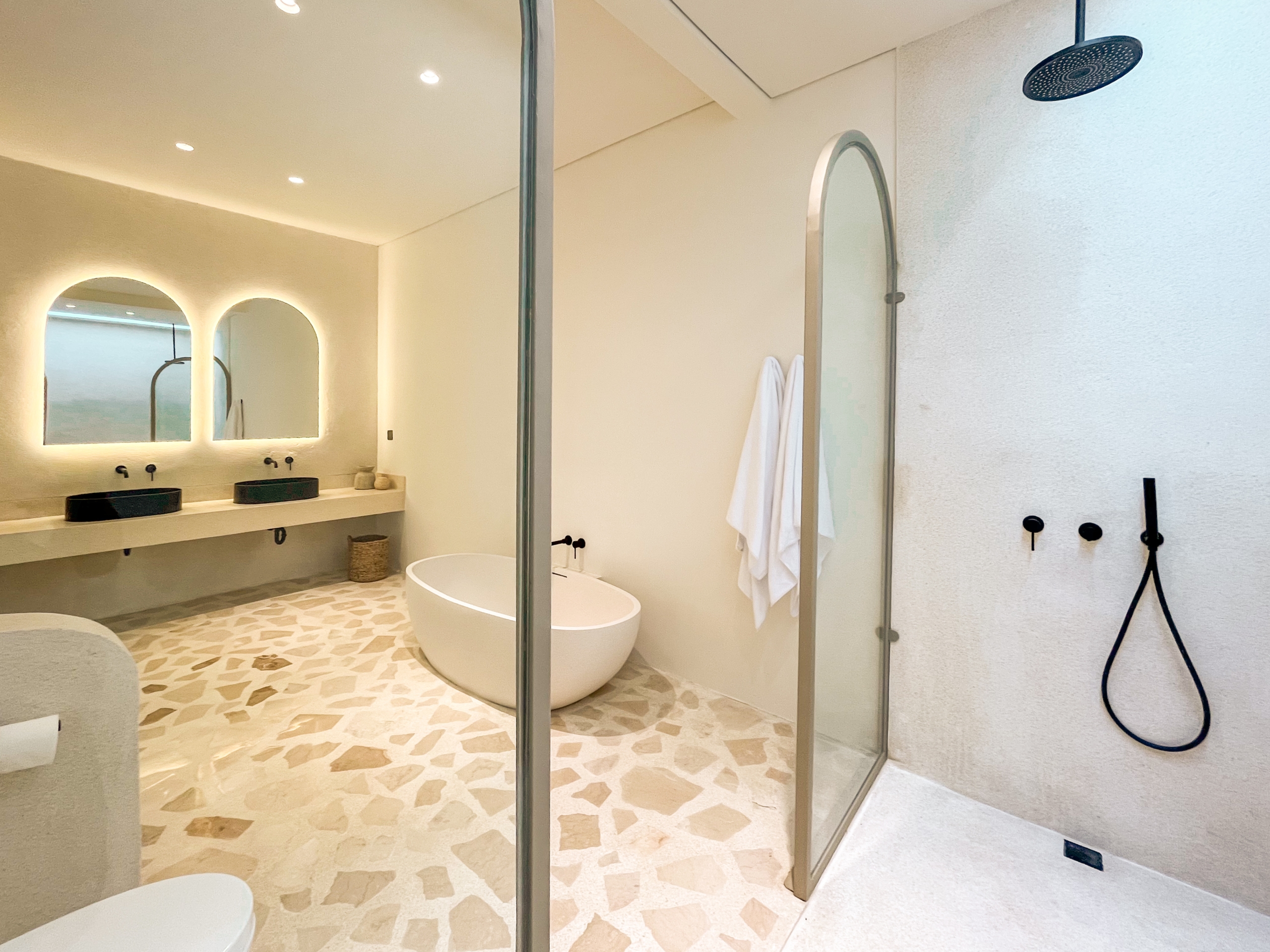 IMG_0500_4669-6a38c7b61fe95 Modern bathroom with a glass shower enclosure, freestanding tub, and double sinks beneath backlit arched mirrors. Decorative pebble floor.