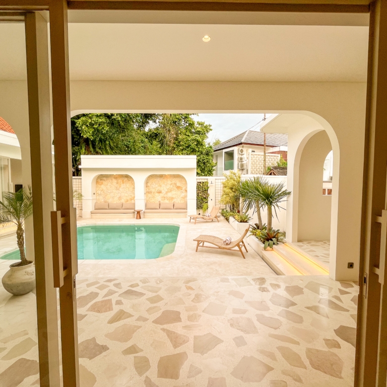 Indoor villa courtyard with a turquoise pool, beige loungers, and greenery framed by white arches and doorways