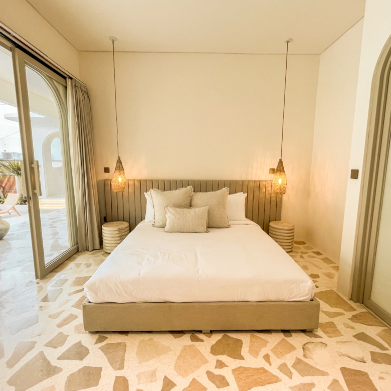 Bedroom with a large bed and beige padded headboard, two woven pendant lights, and neutral decor by a sliding glass door to a balcony/terrace.