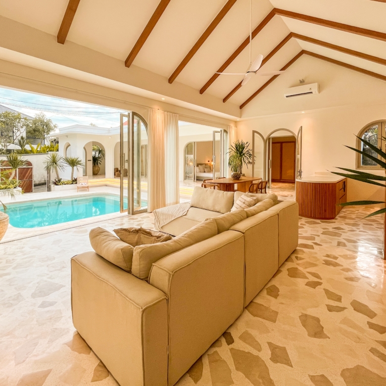 Open-plan living room with vaulted ceiling, dark wood beams, and beige sofa set overlooking a pool through large glass doors of the patio.