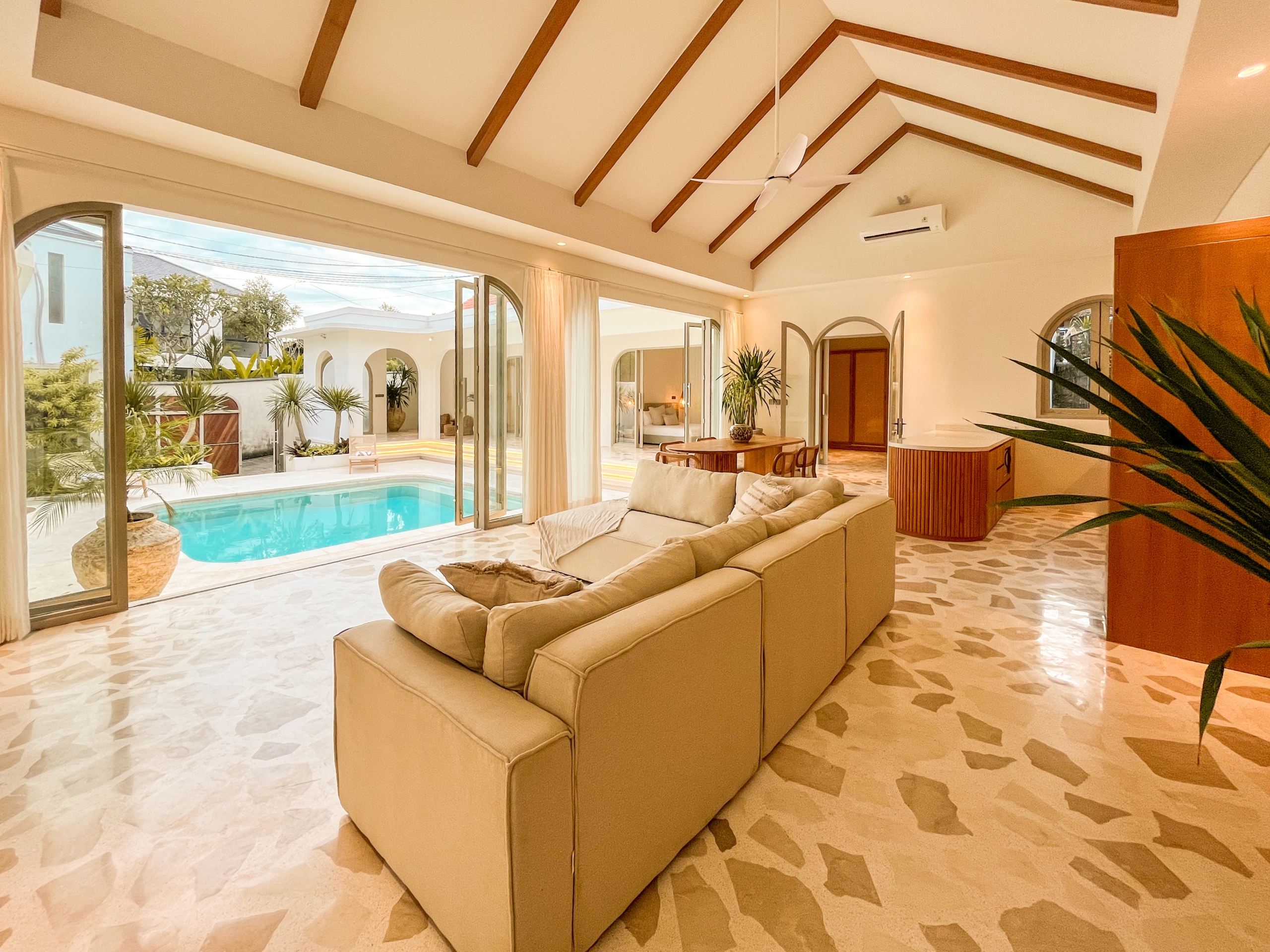 IMG_0578_1__4666-6a38c7b5e2b89 Open-plan living room with vaulted ceiling, dark wood beams, and beige sofa set overlooking a pool through large glass doors of the patio.