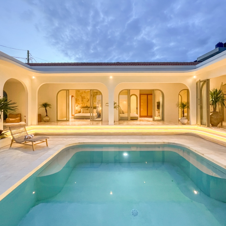 Hotel courtyard at dusk with a rectangular pool, arches, glass doors to a lit interior bedroom, and potted palm plants; warm exterior lighting.