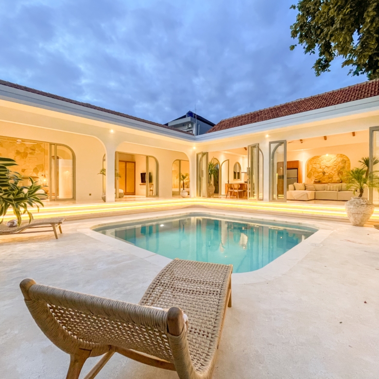 Open-air courtyard with a rectangular pool, white walls, and arches, warm interior lighting, and wicker lounge chair in the foreground at dusk