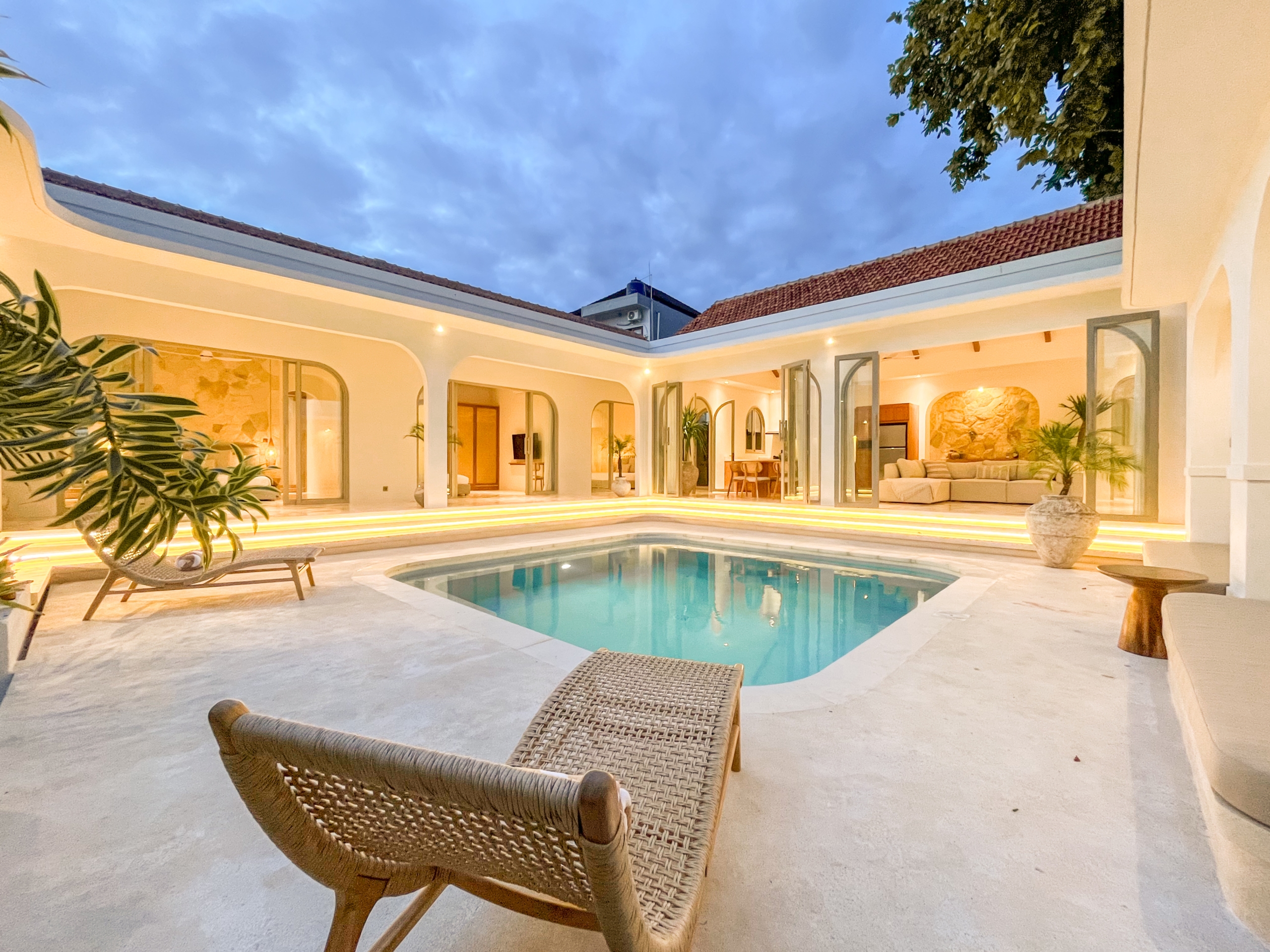 IMG_0589_4710-6a38c7b71fa99 Open-air courtyard with a rectangular pool, white walls, and arches, warm interior lighting, and wicker lounge chair in the foreground at dusk